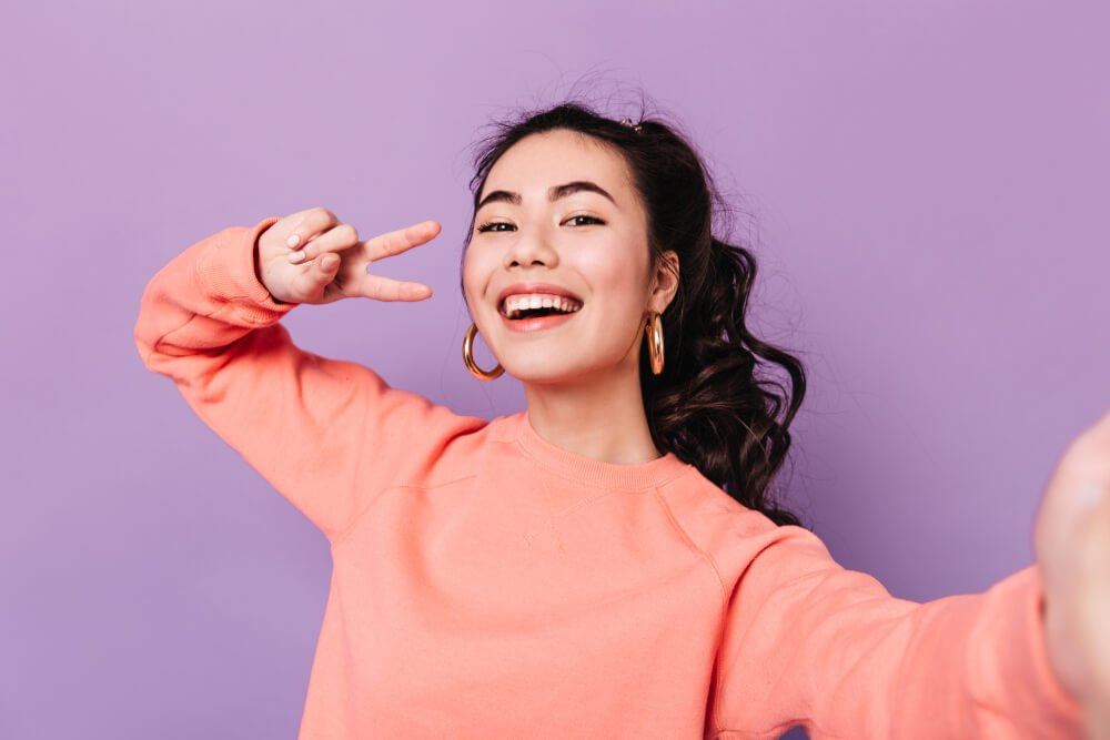 lovely-korean-woman-posing-with-peace-sign-laughing-asian-young-woman-taking-selfie-purple-background (1)